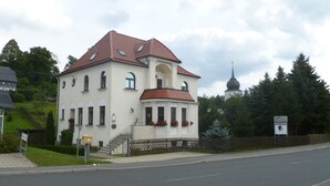 A two-story building with a red-tiled roof, a central entrance with steps, and a small porch with flower boxes. - Pension Wernesgrün (Steinberg)