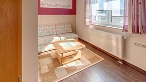 A room with a wooden floor, a sofa, a small table, a radiator, and a window with curtains. - Pension Wernesgrün (Steinberg)