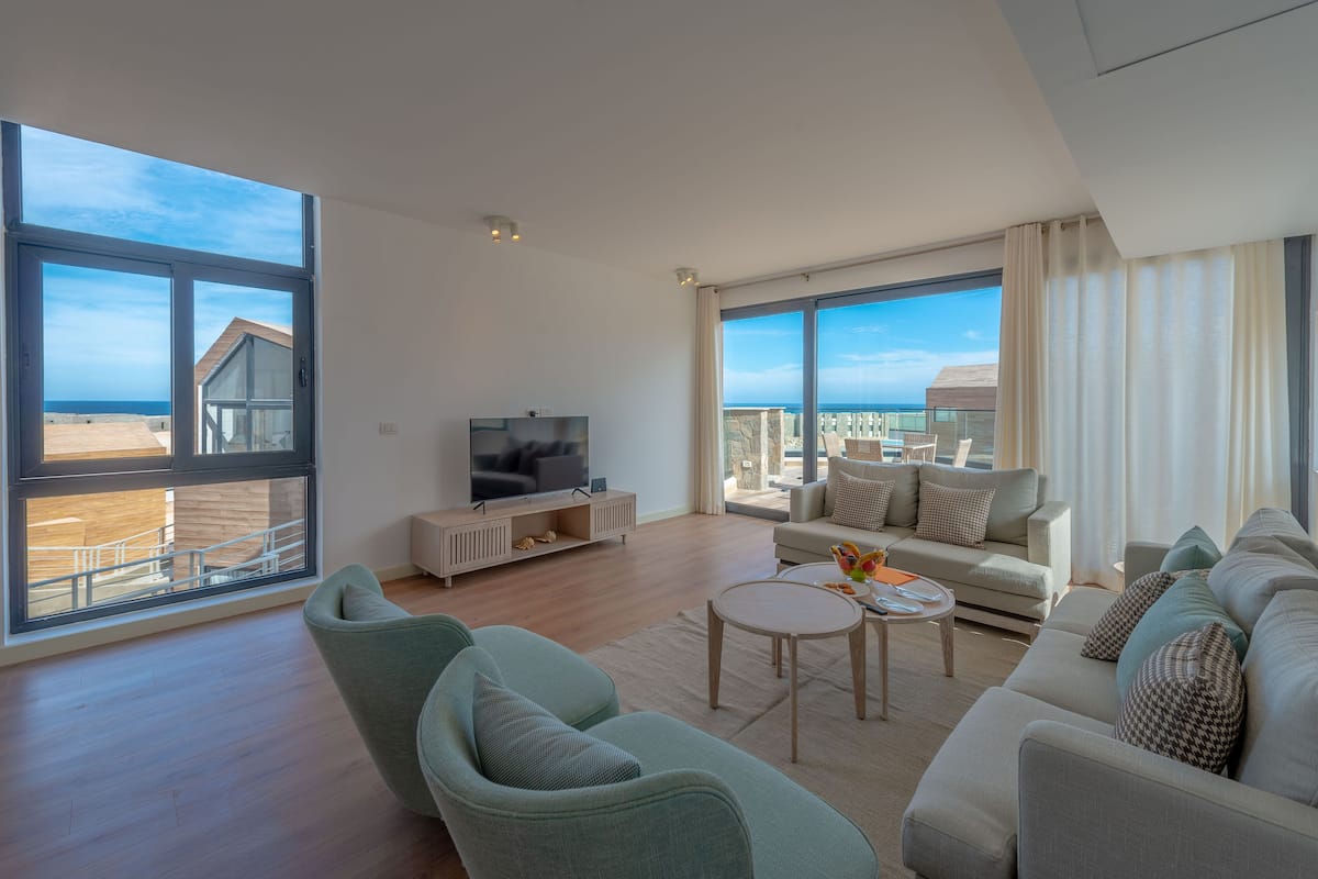 A spacious living room with a large TV, comfortable seating, and a view of the beach.