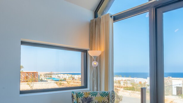 A room with a large window, a patterned armchair, and a view of the sea and buildings. - VESTA - Soma Bay Residence (Soma Bay)