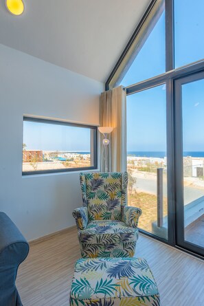 A room with a large window, a patterned armchair, and a view of the sea and buildings. - VESTA - Soma Bay Residence (Soma Bay)