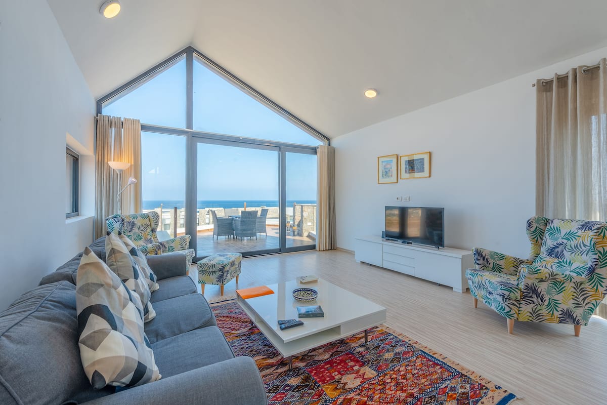 A spacious living room with a large window, a sofa, a coffee table, a TV, and a patterned armchair.