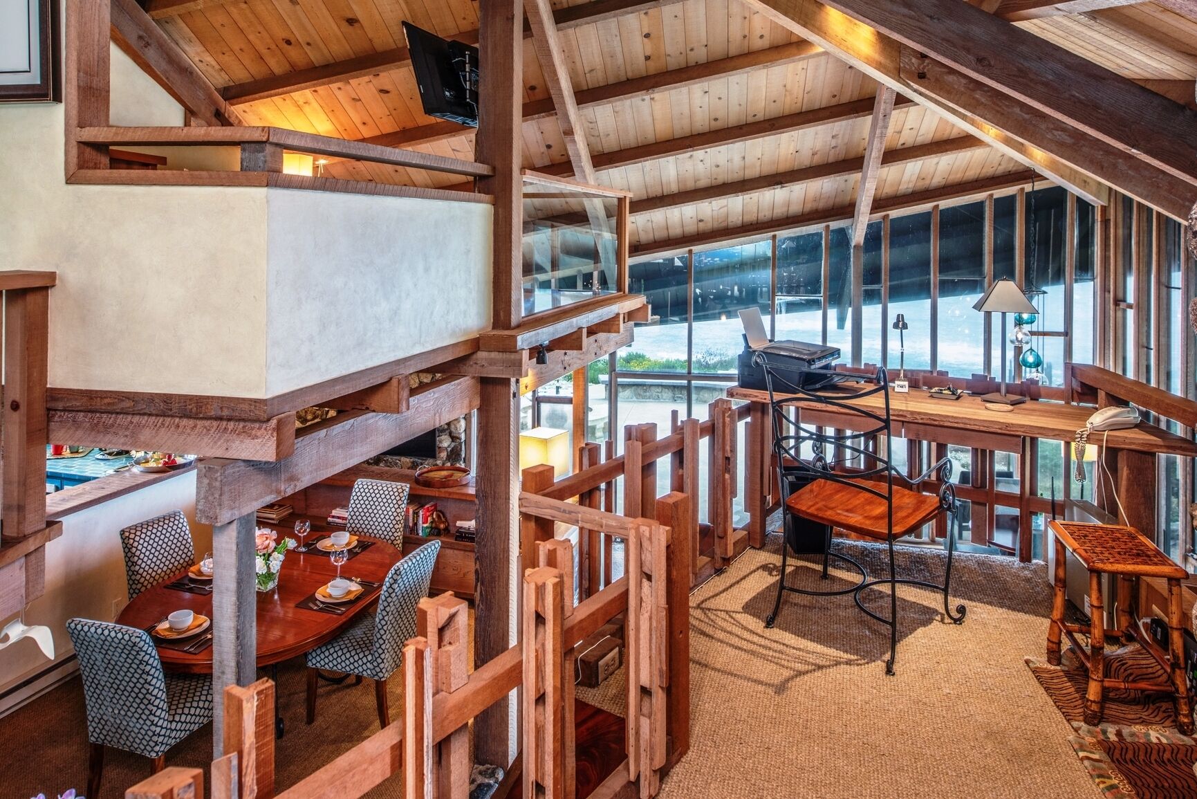 A rustic wooden interior with a dining area, a workspace, and large windows offering views of the outdoors.