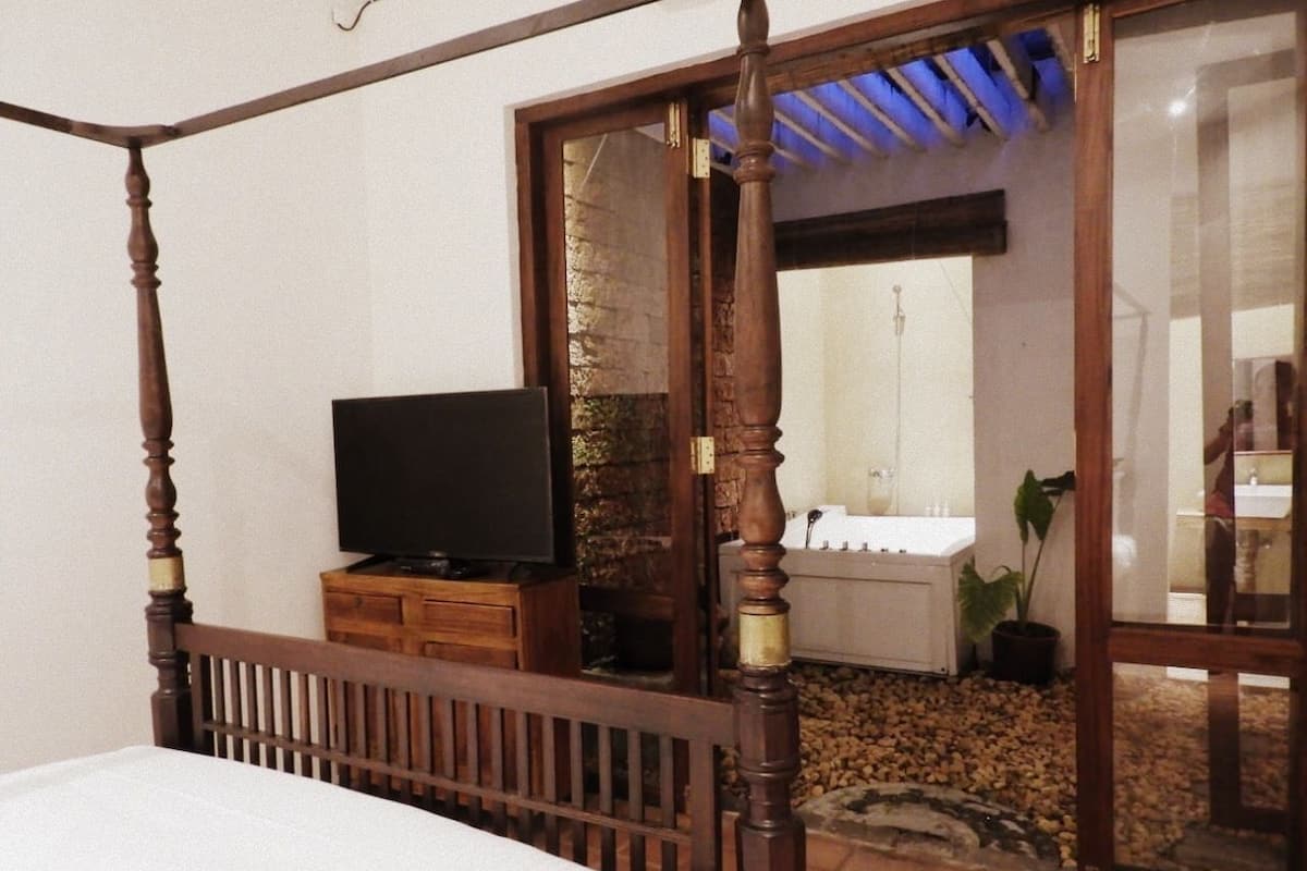 A bedroom with a four-poster bed, a television on a wooden cabinet, and a view of a bathroom through a glass door.
