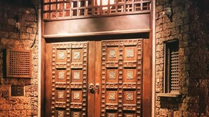 Une grande porte à deux battants en bois, ornée de sculptures complexes, intégrée à un bâtiment en pierre dont l’entrée est pavée de briques.