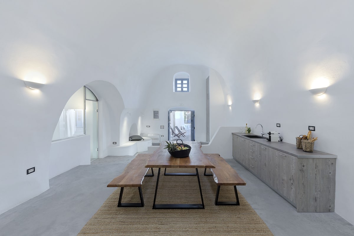 A modern kitchen with a wooden table and bench, a built-in island, and a view through an arched doorway.