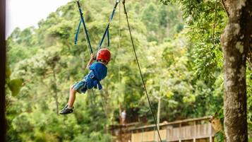 A person on a zip line in a forested area with a wooden bridge in the background.