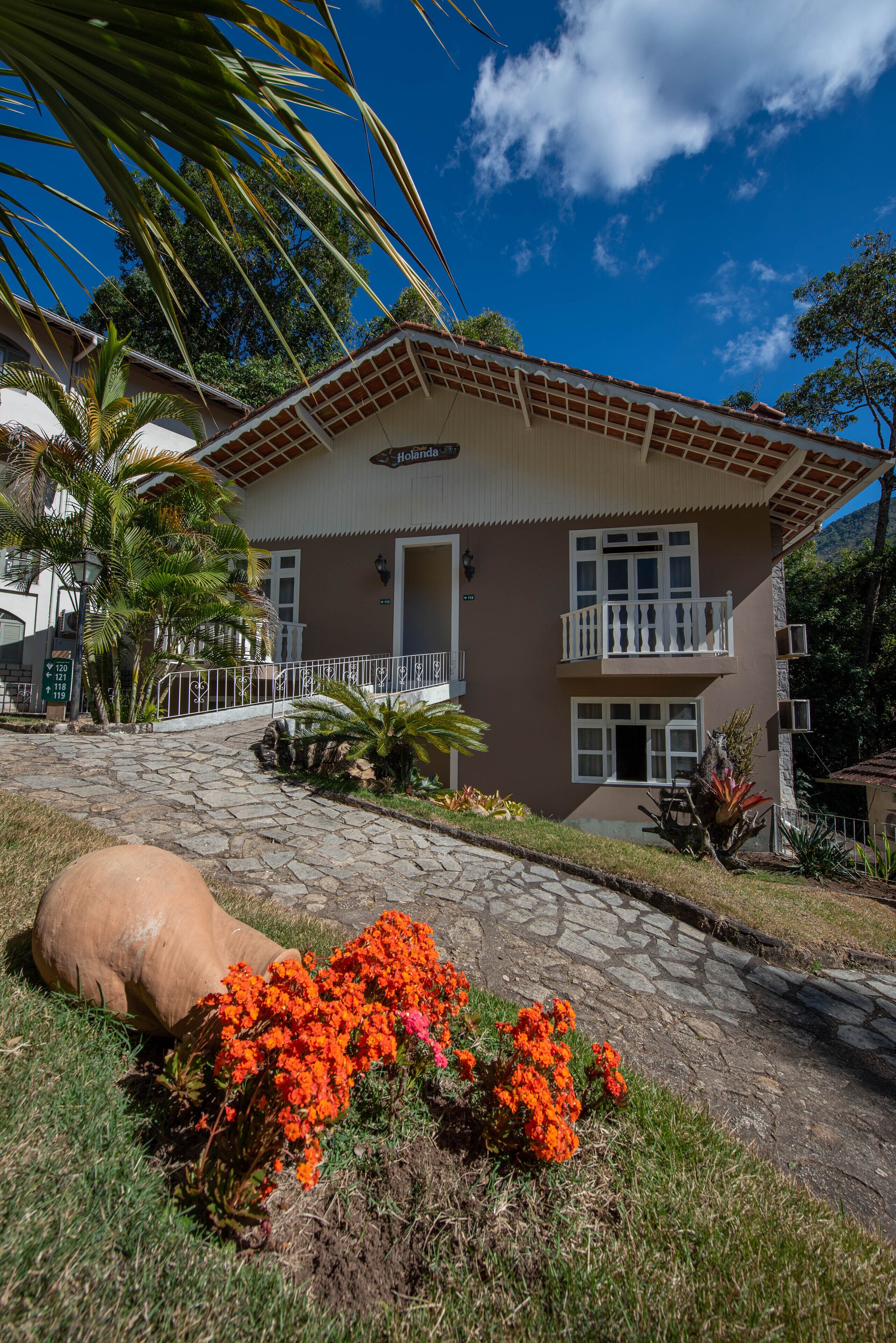 A two-story building with a balcony, a stone pathway leading to the entrance, and a tropical garden with orange flowers.