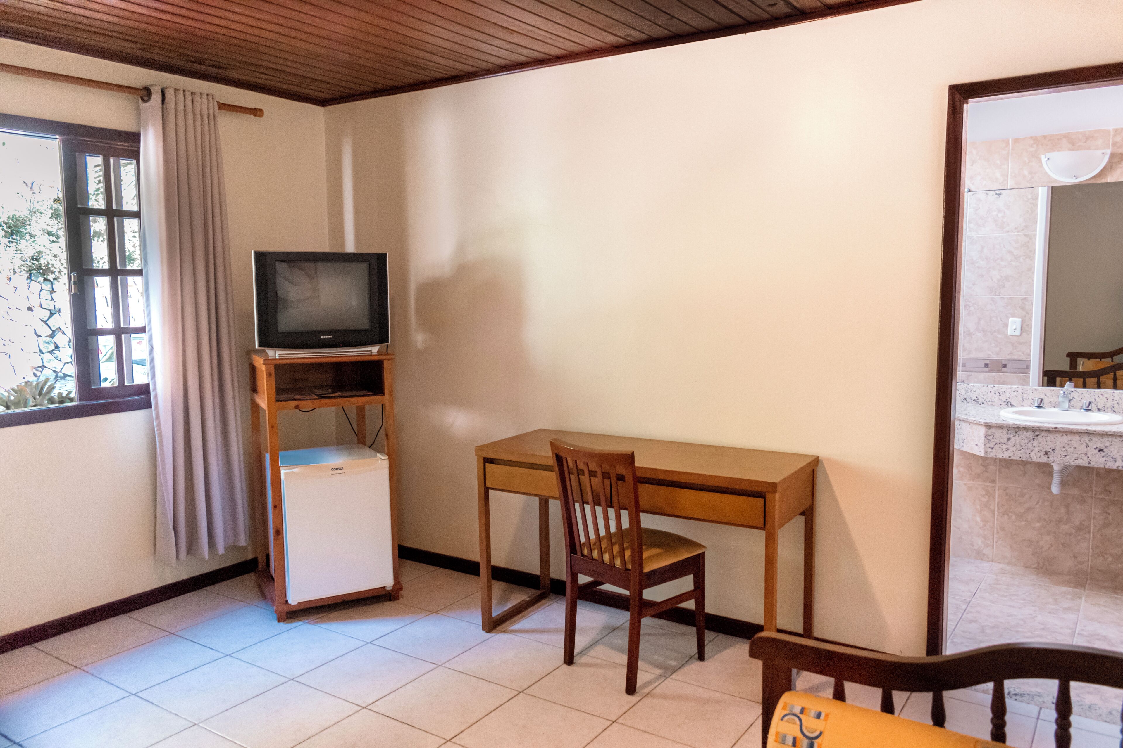 Una habitación con un escritorio de madera, un televisor sobre un soporte, un refrigerador pequeño y una silla.