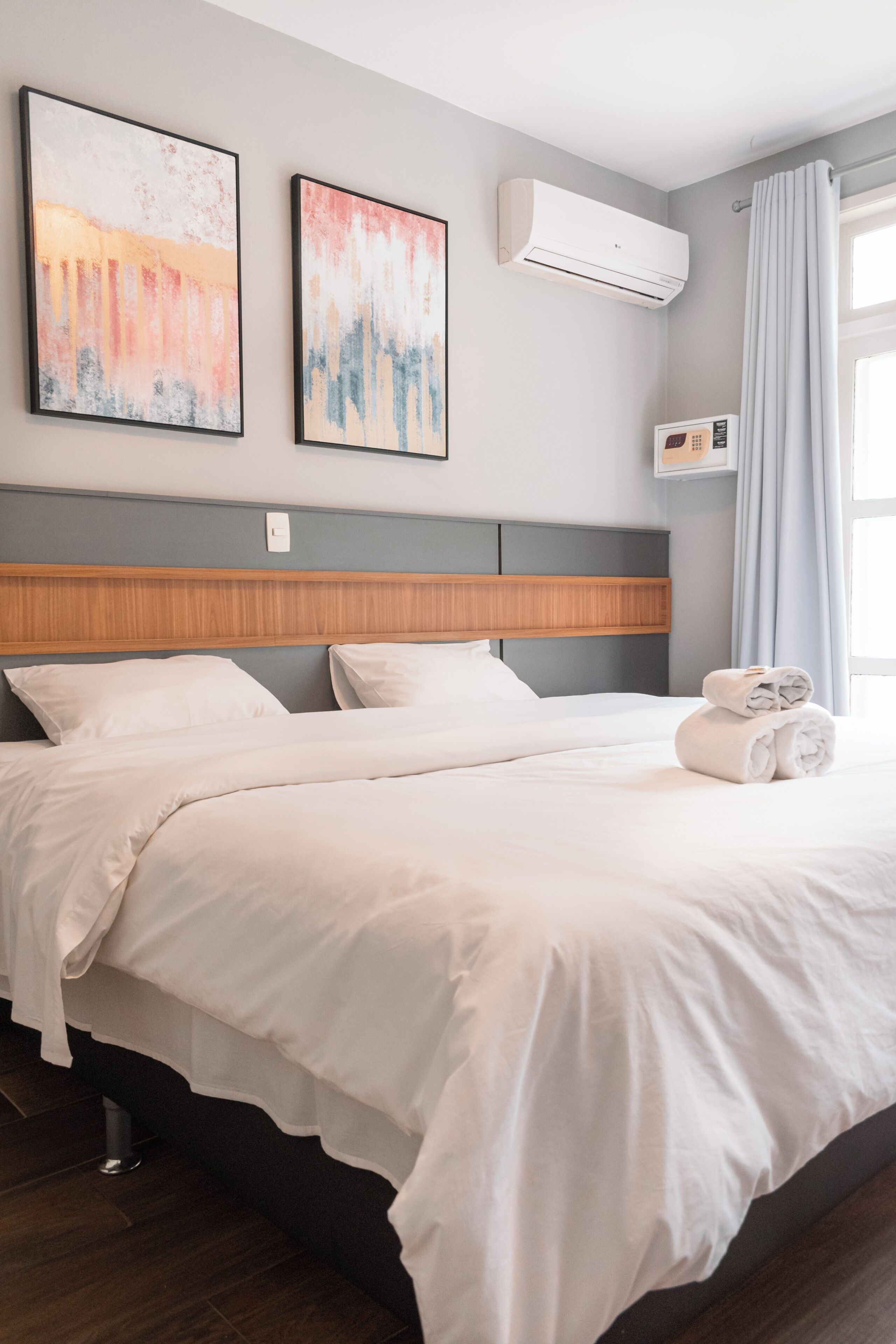 A hotel room with a bed, two wall-mounted air conditioners, and a window with curtains.