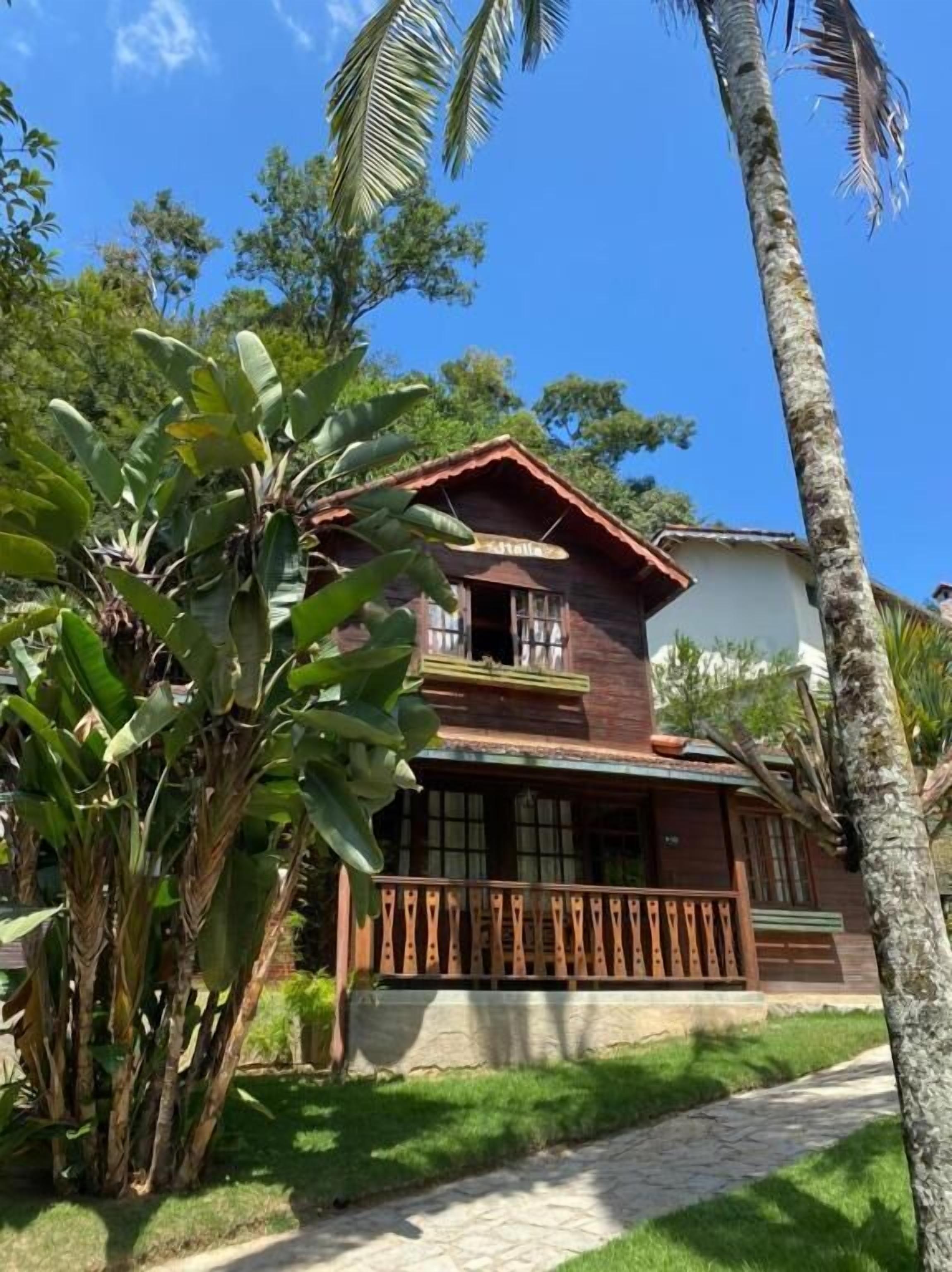 Uma casa de madeira de dois andares com uma varanda, cercada por vegetação exuberante e palmeiras.
