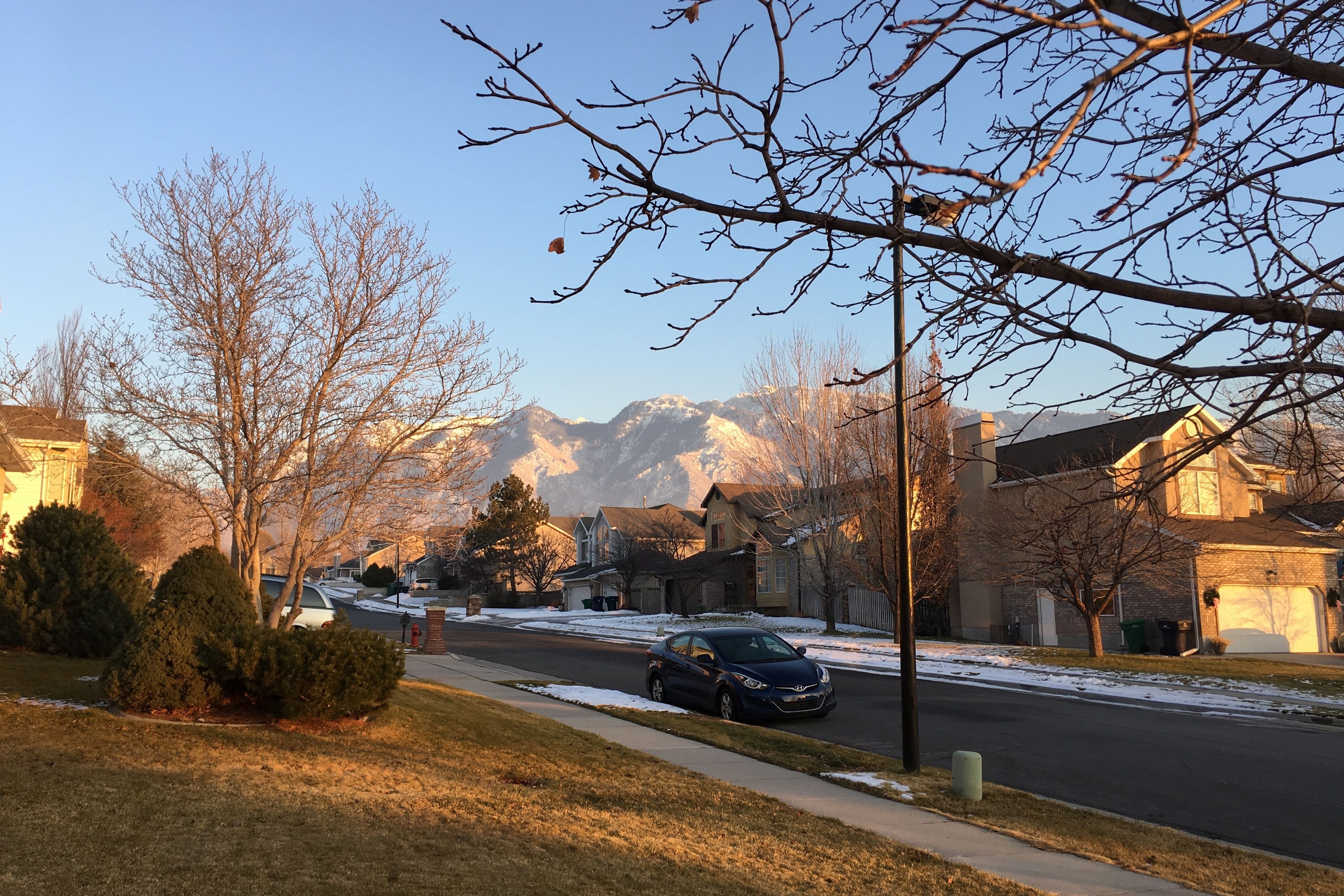 Une rue résidentielle bordée de maisons, avec une voiture garée et des montagnes en arrière-plan.