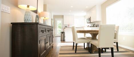 A modern kitchen with a dining area, featuring a dark wooden cabinet, white chairs, and a striped rug.