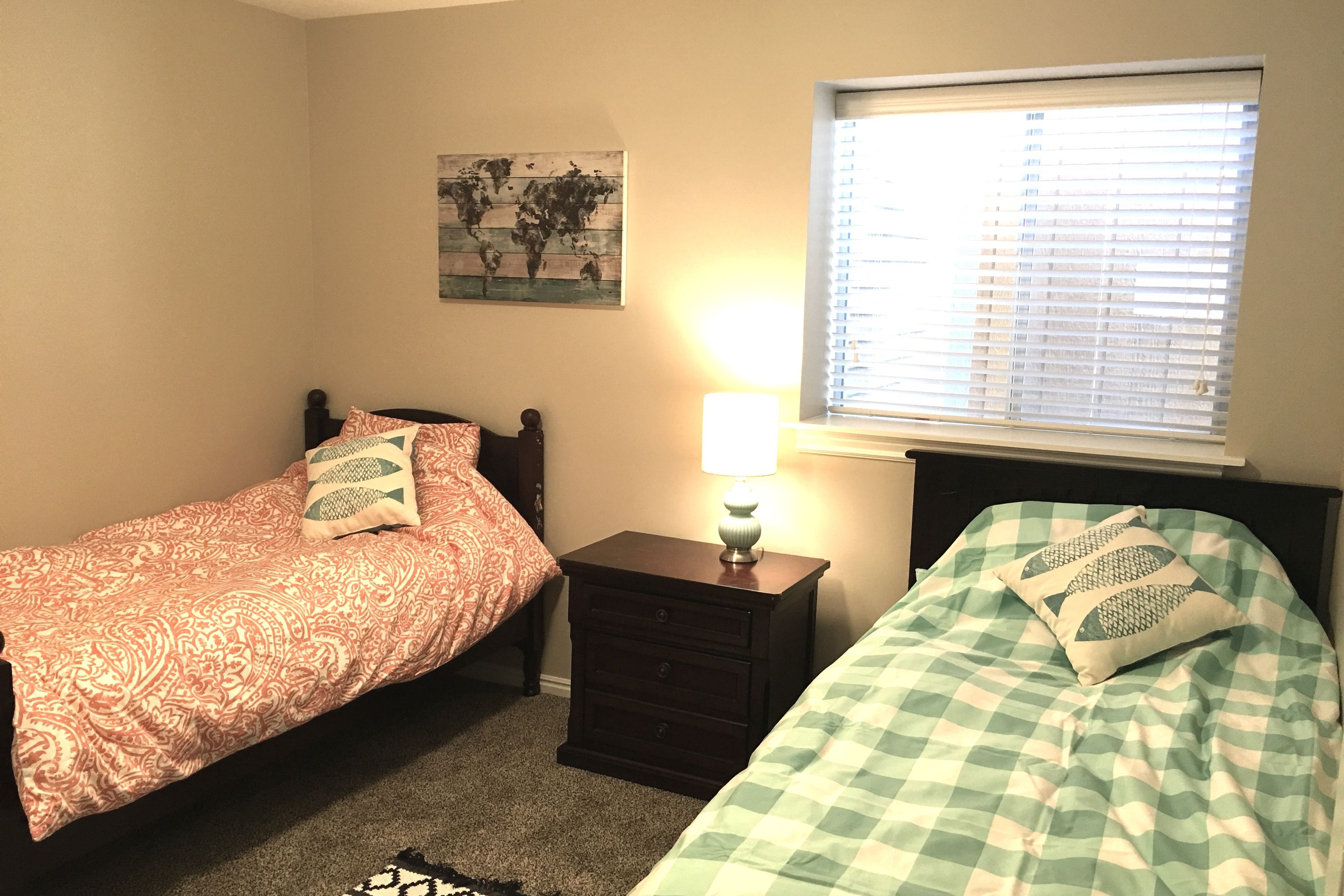 A bedroom with two beds, a nightstand with a lamp, a world map on the wall, and a window with blinds.