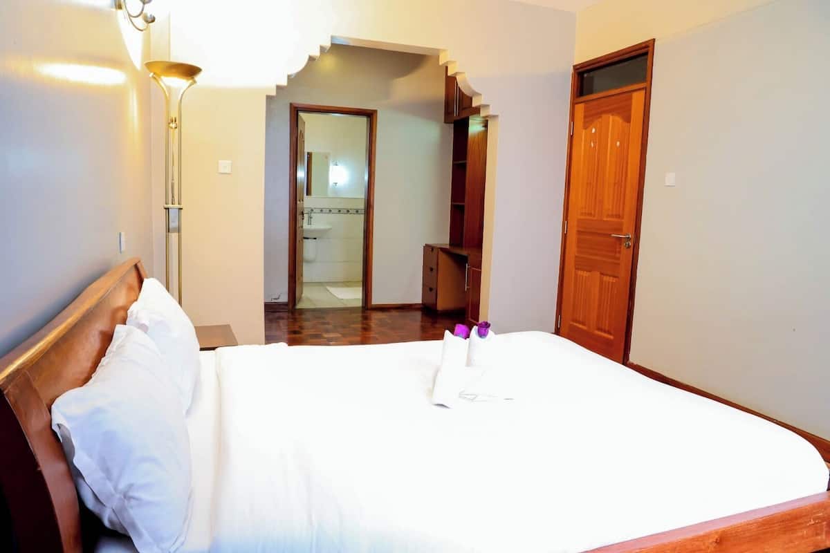 A hotel room with a bed, a nightstand, a lamp, a wardrobe, and a bathroom visible through an open door.