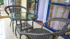 A glass-top table with wicker chairs, a blue railing, and a view through large windows. - KenGen Furnished and Serviced Apartments (Nairobi)