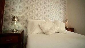 A neatly made bed with white linens and pillows, a wooden nightstand with a lamp, and a wall with a geometric pattern. - Villa Cristal Holguin (Holguín)