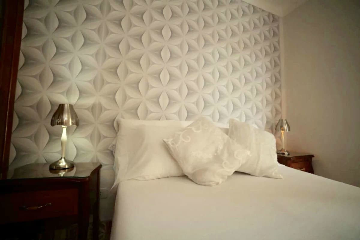 A neatly made bed with white linens and pillows, a wooden nightstand with a lamp, and a wall with a geometric pattern.