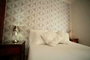 A neatly made bed with white linens and pillows, a wooden nightstand with a lamp, and a wall with a geometric pattern. - Villa Cristal Holguin (Holguín)