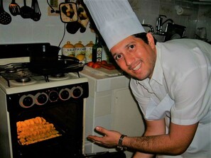A chef in a white uniform and hat standing next to an oven with a tray of food inside. - Villa Cristal Holguin (Holguín)