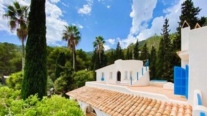 A white building with blue accents, a tiled roof, and a view of surrounding greenery and trees.