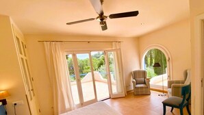 A bedroom with a bed, two chairs, a ceiling fan, and a view through the glass doors.