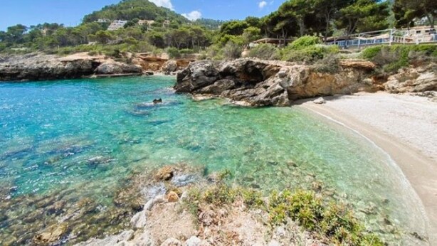 A coastal beach with clear turquoise water, rocky shorelines, and a few buildings in the background.