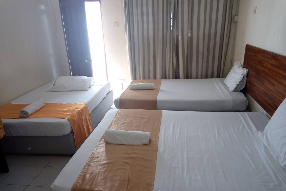 Una habitación de hotel con dos camas individuales, cada una con un colchón blanco y un cabecero marrón. Hay una ventana con cortinas y el suelo es de baldosas.