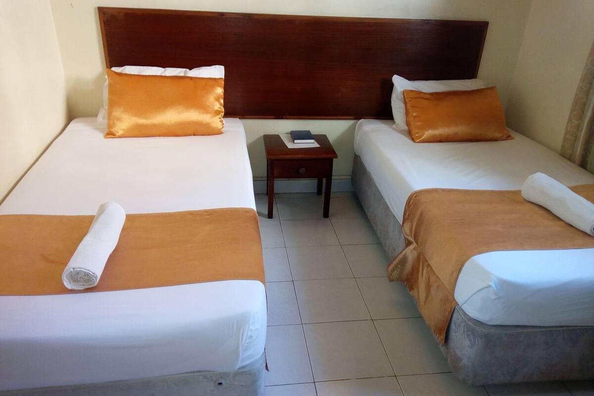 Habitación de hotel con dos camas individuales, cabecero de madera, una mesita pequeña y suelo de baldosas.