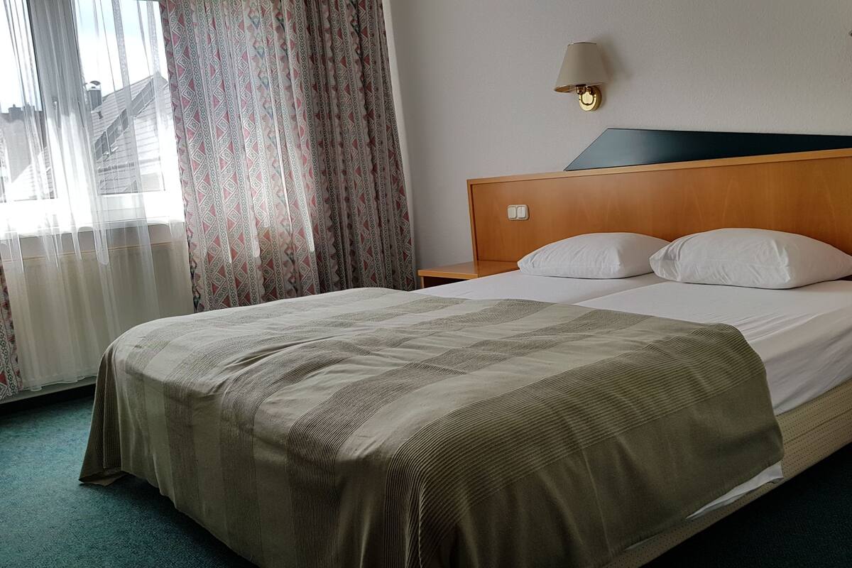 Habitació d'hotel amb un llit gran, un reposacapses de fusta, dues làmpades de sobretaula i una finestra amb cortines estampades.