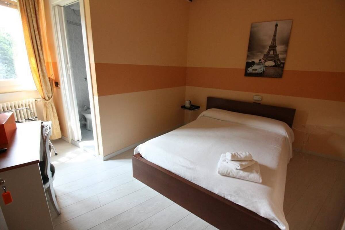 Une chambre d’hôtel avec un lit, une table de chevet, une fenêtre avec des rideaux, un radiateur et un tableau représentant la Tour Eiffel.