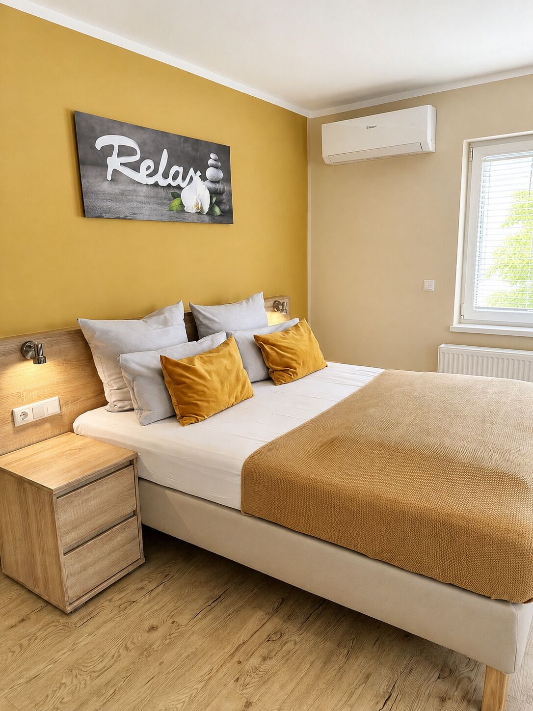 A bedroom with a bed, bedside tables, a wall-mounted air conditioner, and a window with blinds.