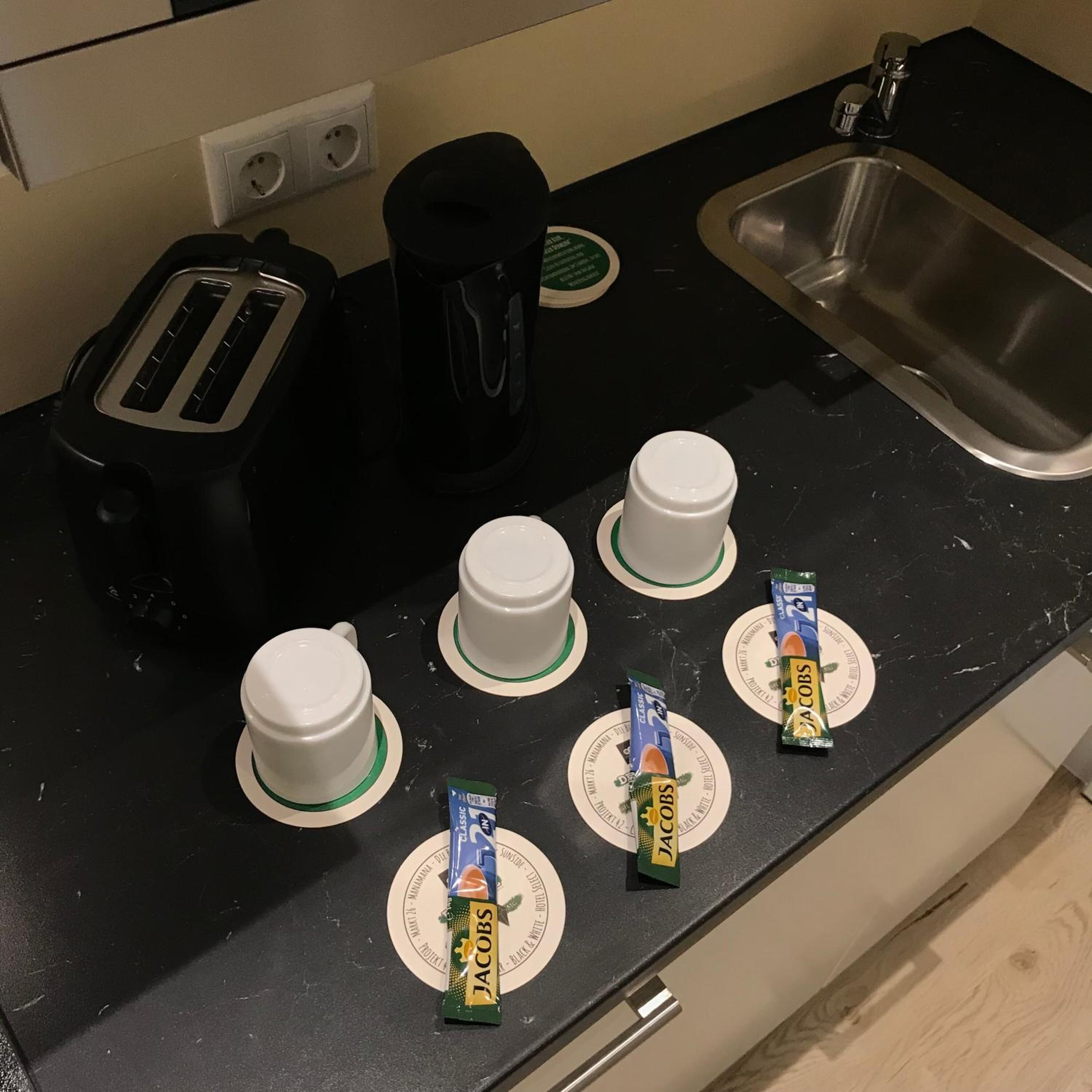 Un plan de travail de cuisine avec un grille-pain, une machine à café, un évier et quatre tasses avec des couvercles. On y trouve également deux paquets de café Jacobs.
