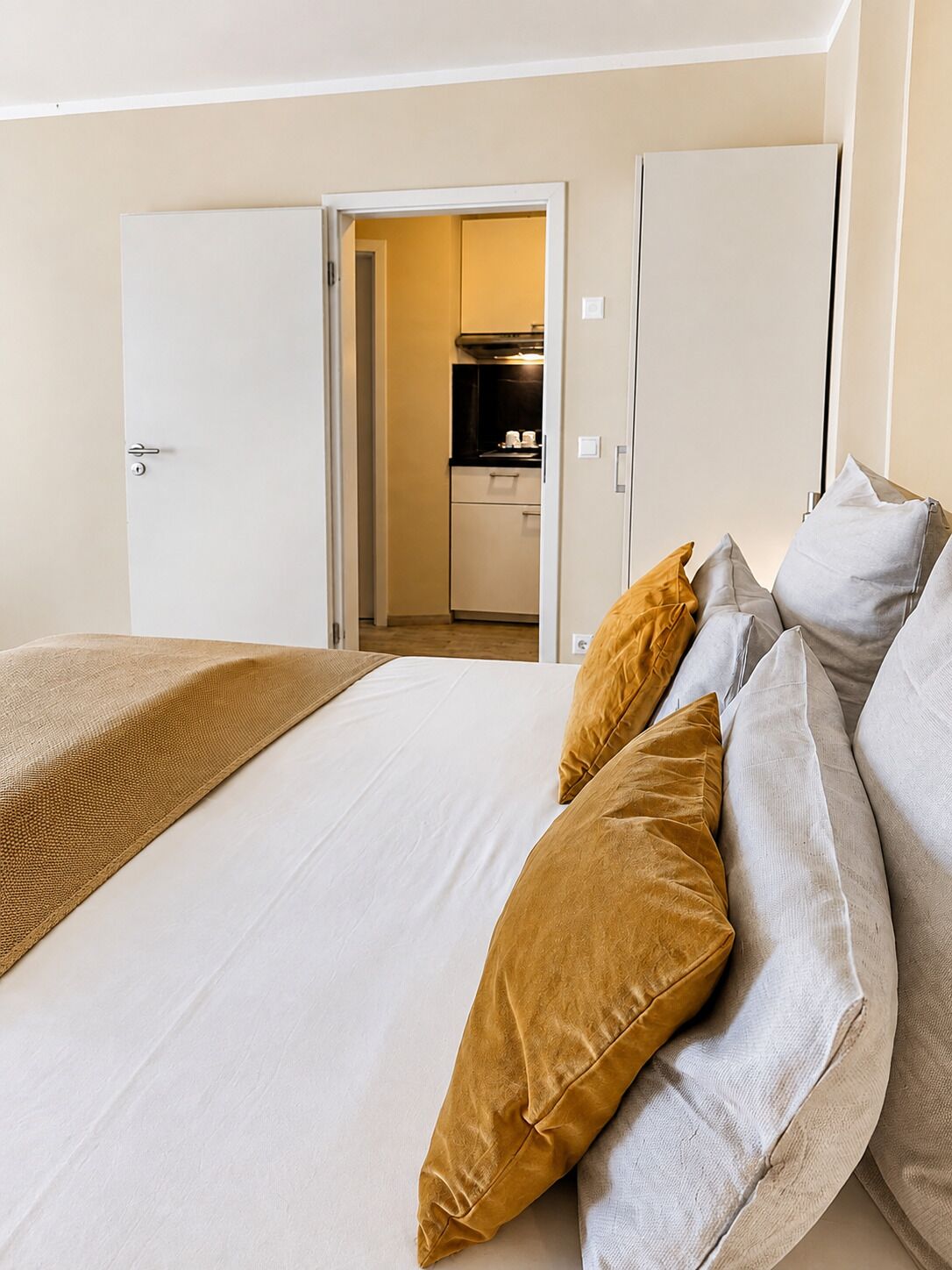 Une chambre d’hôtel avec un lit, des oreillers et une vue sur une cuisine, visible à travers une porte ouverte.