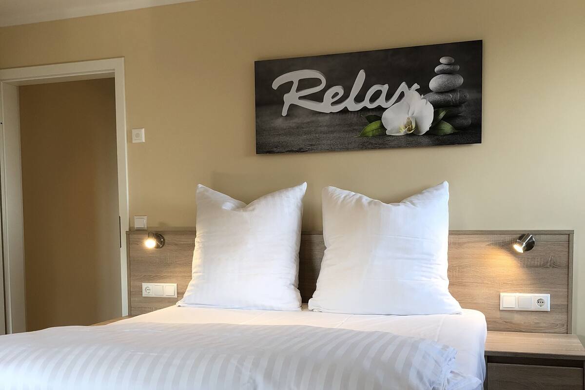 A neatly made bed with white linens, a headboard with a 'Relax' sign, bedside lamps, and a wooden nightstand.