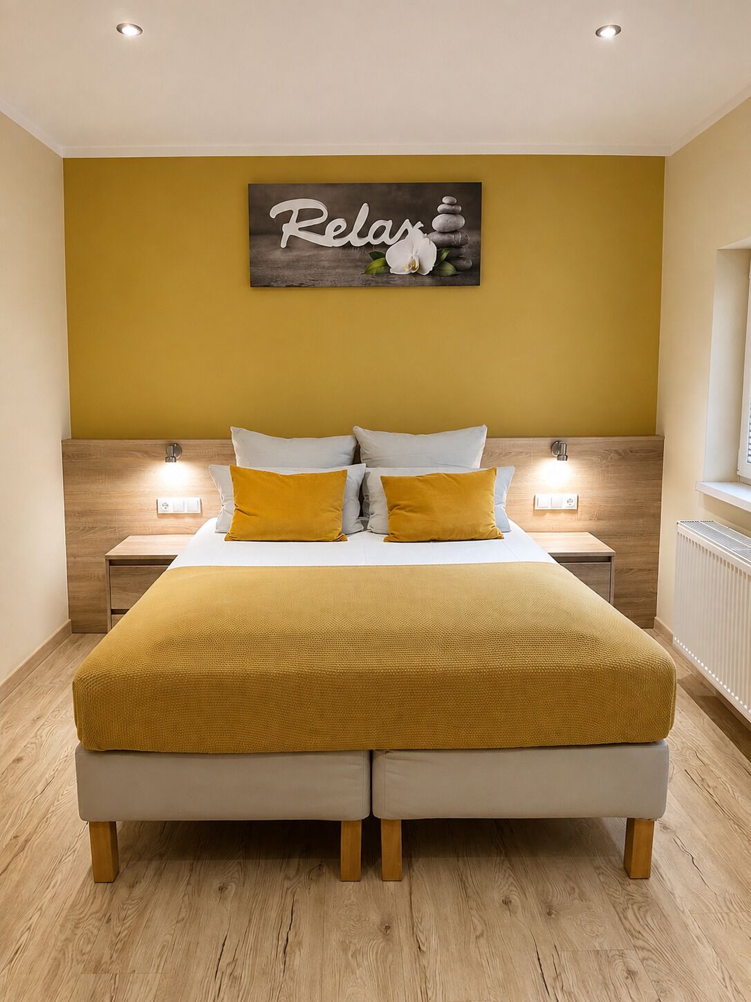 A double bed with a mustard-colored blanket and pillows, two bedside lamps, a wall-mounted artwork with the word 'Relax' and a stack of stones, and a wooden headboard.