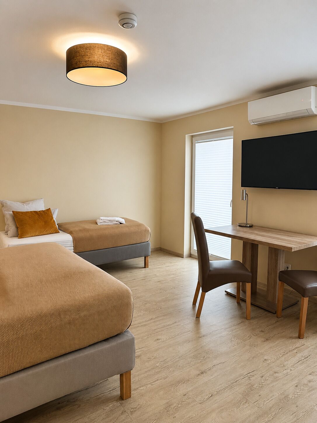 A hotel room with two beds, a desk with a chair, a flat-screen TV, and an air conditioning unit.