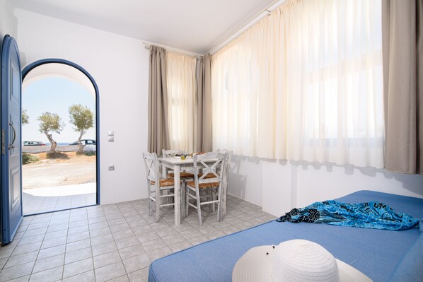 A room with a bed, a dining table with chairs, a view through an open door, and a window with curtains. - Villa George Santorini (Santorini)
