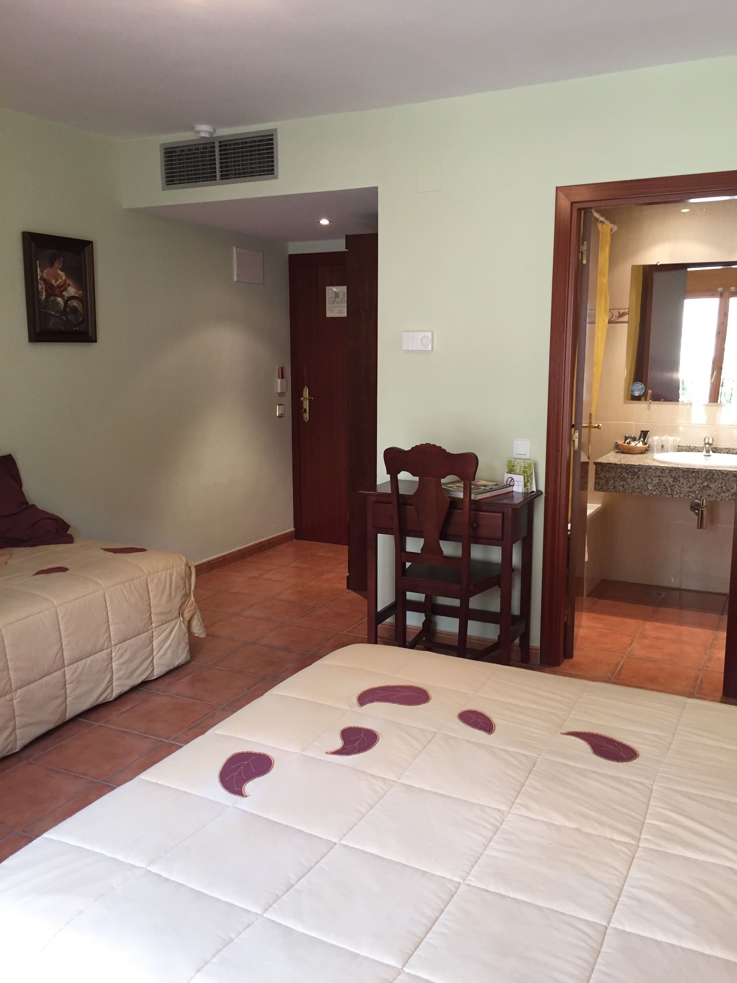 A hotel room with a bed, a desk, a chair, a bathroom with a sink and mirror, and a door leading to another room.