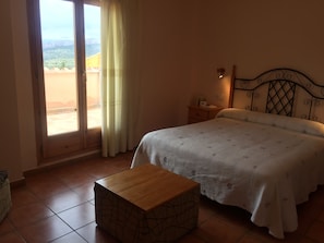 A bedroom with a bed, a wooden nightstand, a bedside table, a window with curtains, and a view of the outdoors. - Hotel Castellote (Castellote)