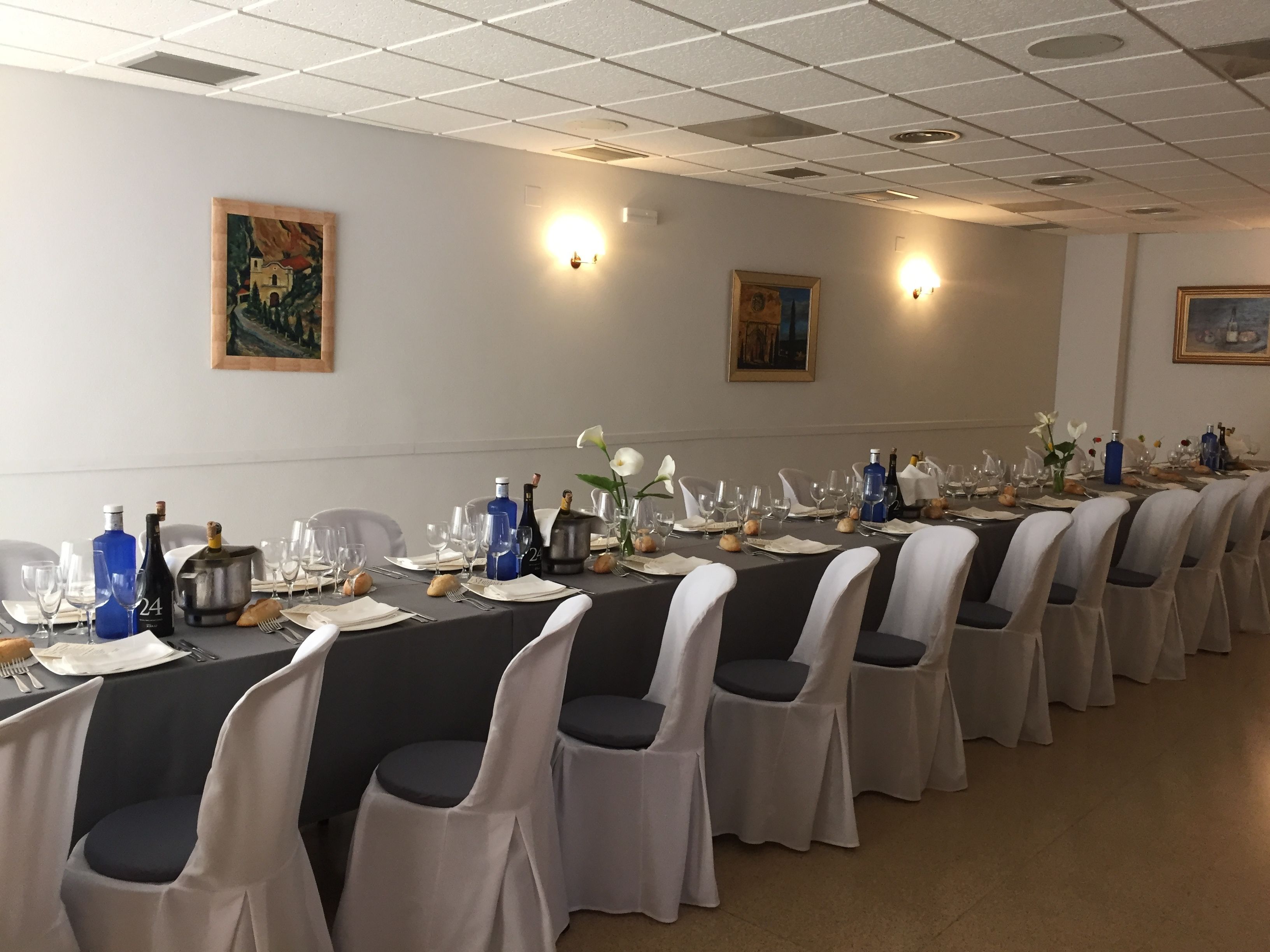 A long dining table set with chairs, wine bottles, and glasses in a well-lit room with ceiling lights and wall-mounted lamps.