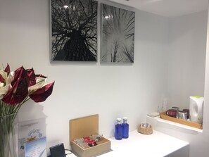 Ett rum med ett vitt skrivbord, en vas med röda och vita blommor, två inramade bilder av träd, en klocka och en liten hylla med muggar och flaskor.