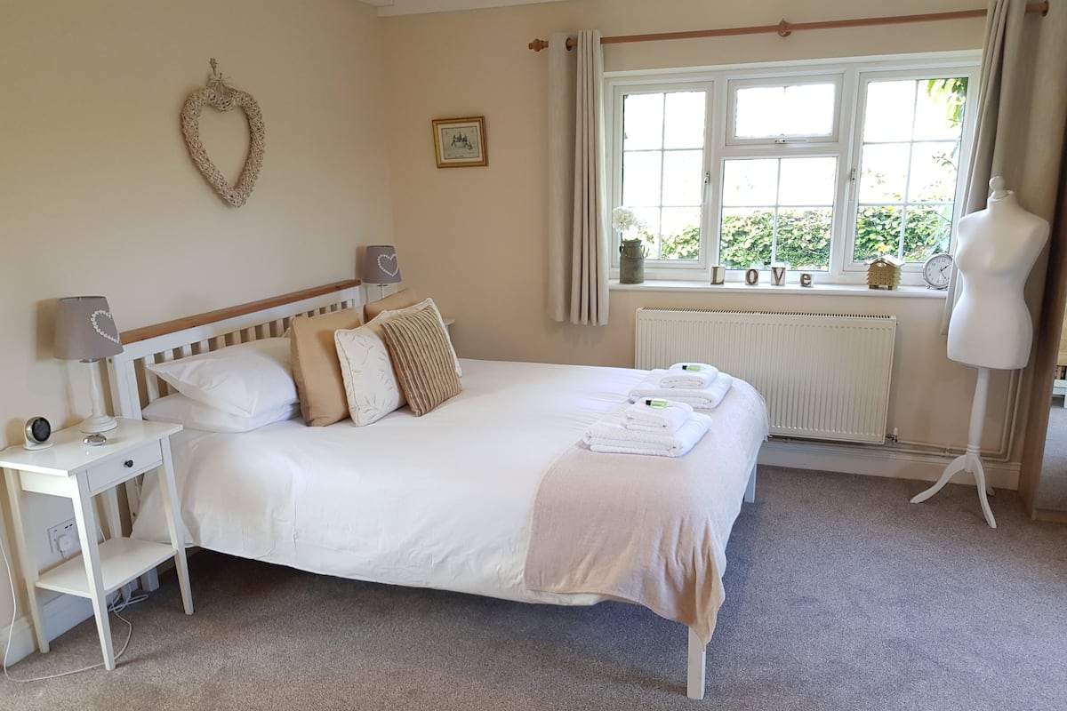 Een net opgemaakt bed met witte lakens en een beige plaid, een nachtkastje met een lamp, een dressoir, en een etalagepop in een kamer met een groot raam en een radiator.