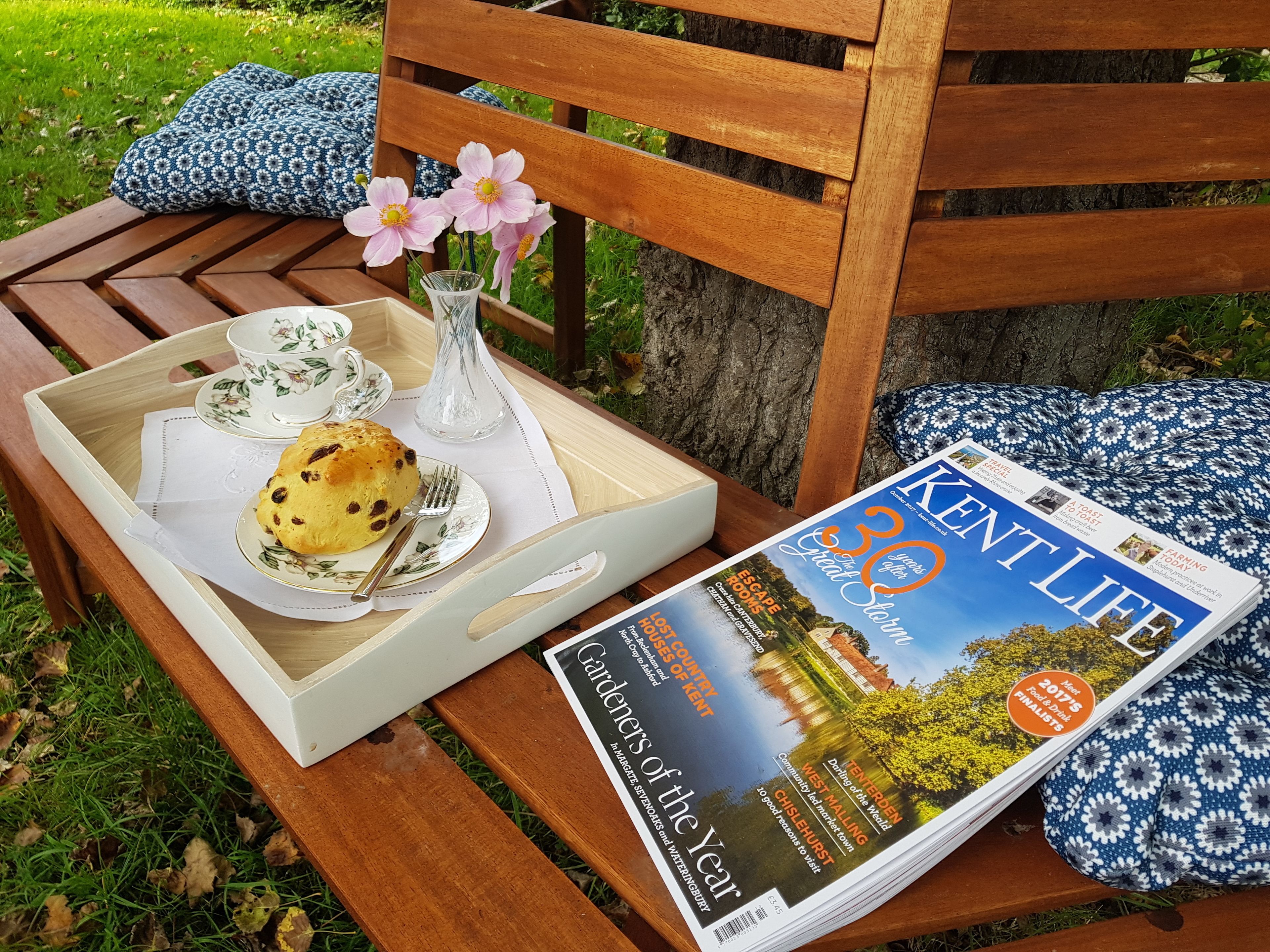 A wooden bench with a tray containing a scone, a teacup, saucer, and a vase with flowers. A magazine titled "Kent Life" is also visible.