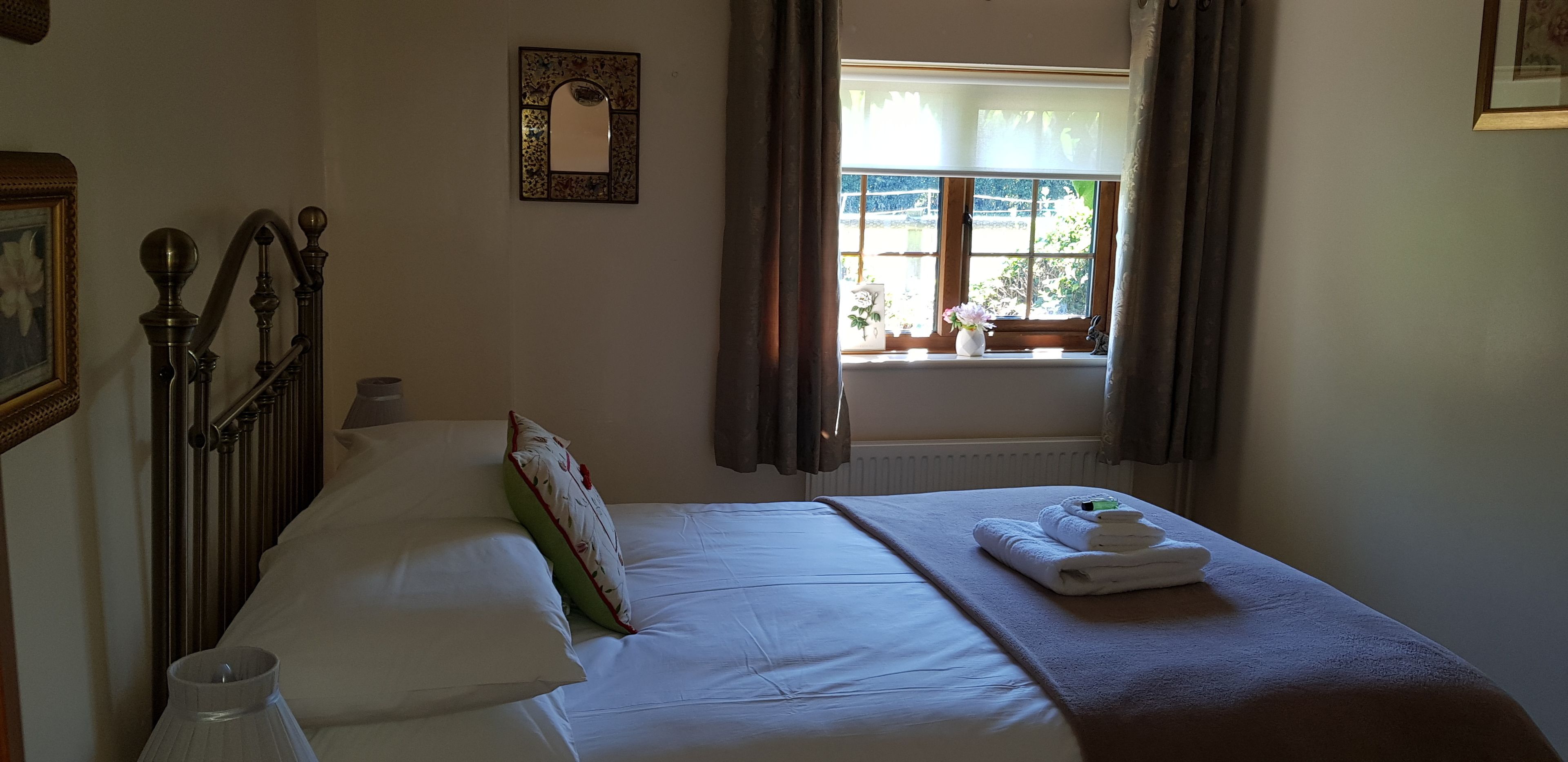 A neatly made bed with white linens and a brown throw blanket, a bedside table with a lamp, a window with curtains, and a framed picture on the wall.