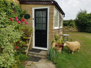 A quaint brick house with a black door, a small porch with a table and chairs, and a garden with a sheep. - Hawthorn Farm Cottages (Canterbury)