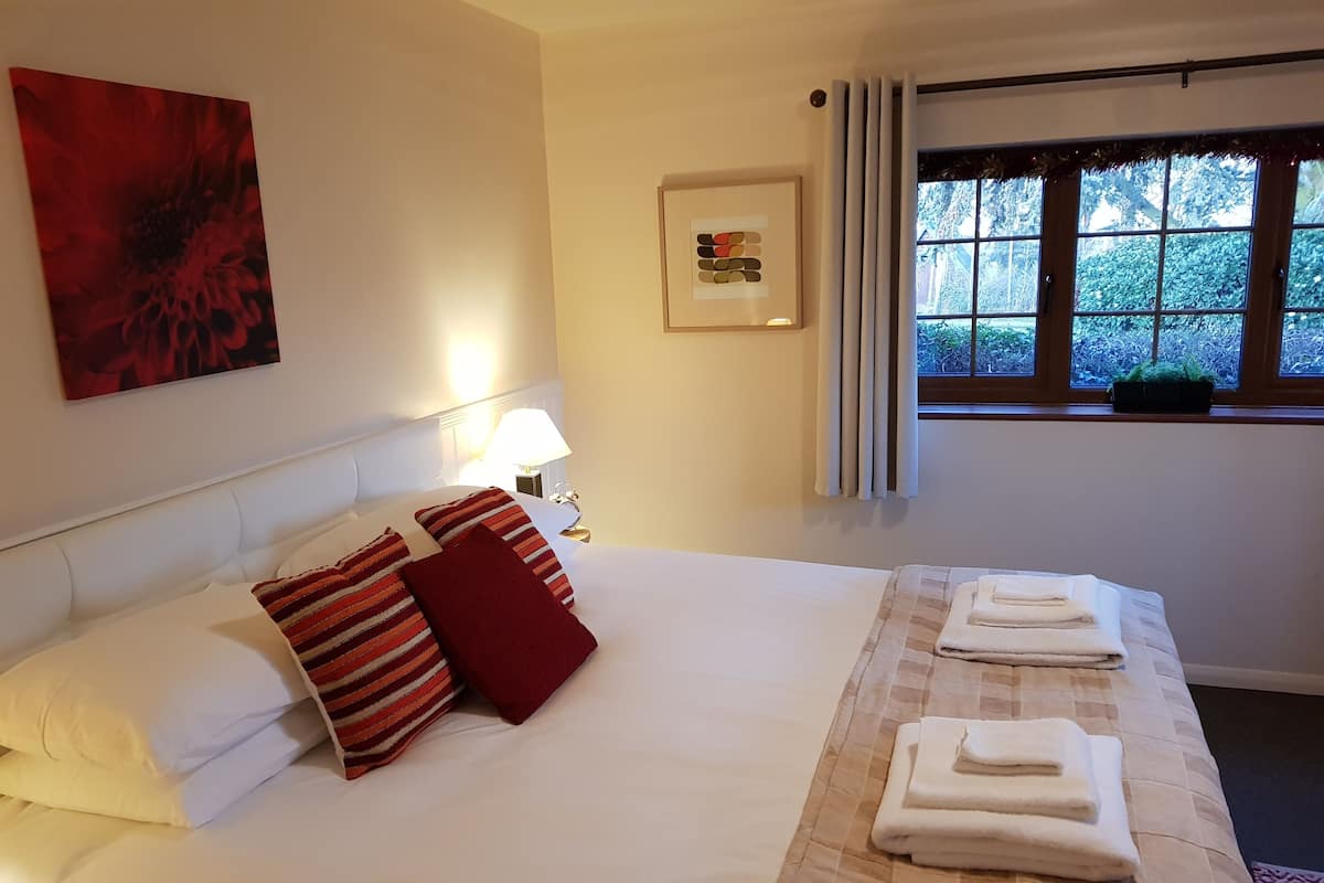 Een hotelkamer met een groot bed, witte beddengoed, een rood kussen, een bedlampje, een raam met gordijnen en een ingelijste foto aan de muur.