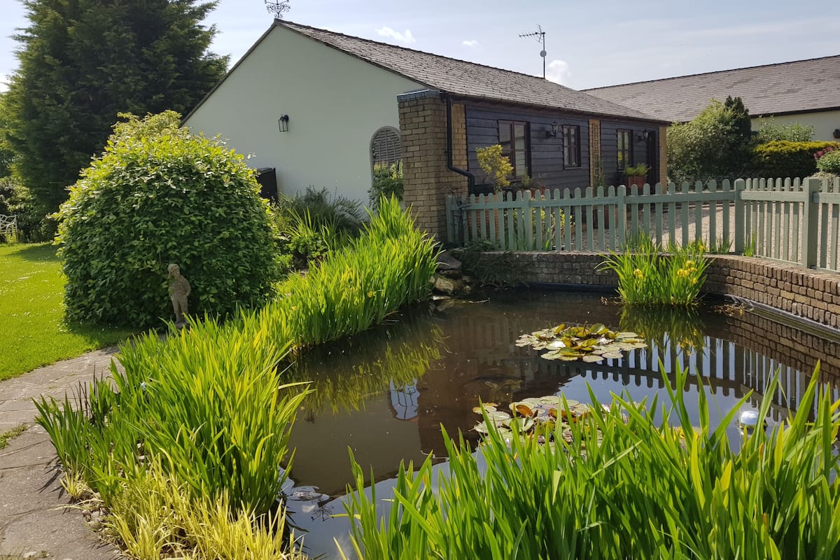 Een vijver met waterlelies, omgeven door weelderige begroeiing en een wit gebouw met een bakstenen uitbouw.