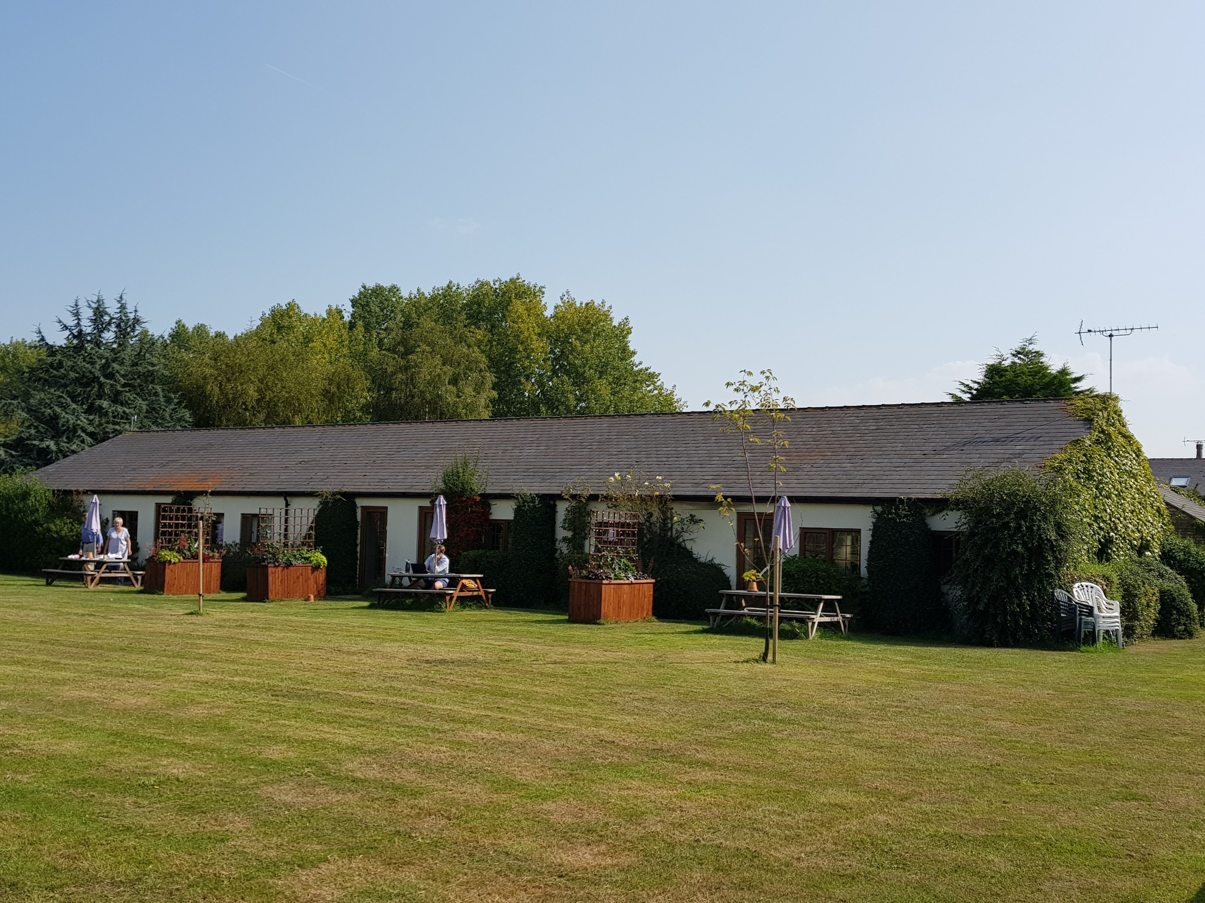 Een gebouw met een plat dak, omgeven door een gazon en bomen. Er zijn terrassen met tafels en stoelen.