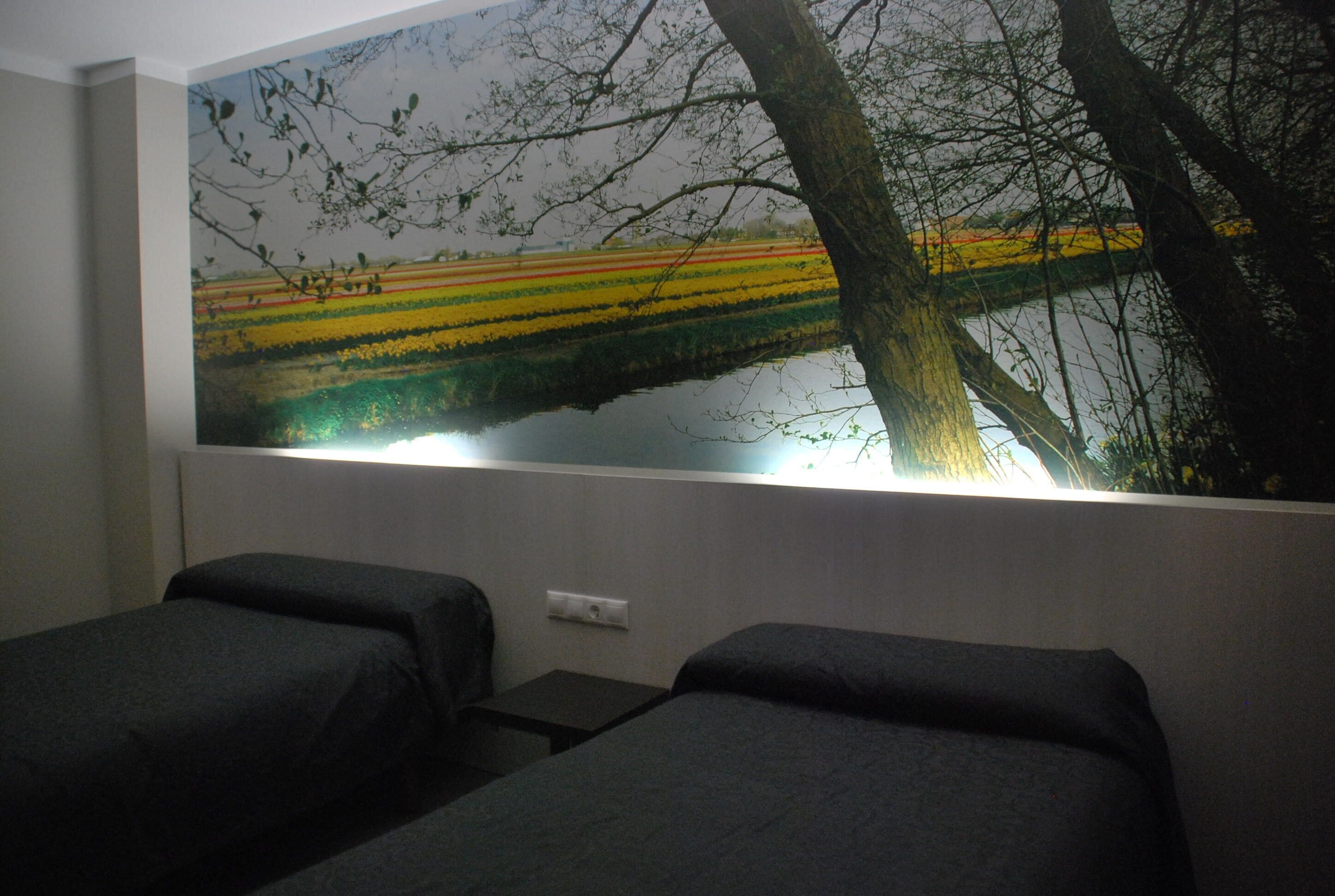 Une chambre d’hôtel avec deux lits simples, une grande fenêtre ornée d’une fresque représentant un paysage et une table de chevet.